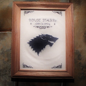 May include: A framed cross-stitch featuring the House Stark sigil, a direwolf, with the text "House Stark, Winter is Coming".