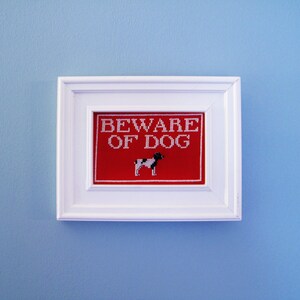 以下が含まれることがあります： 赤い背景に白い文字で「Beware of Dog」と書かれた白い額縁の看板。文字の下には、小さく黒と白の犬のシルエットがあります。