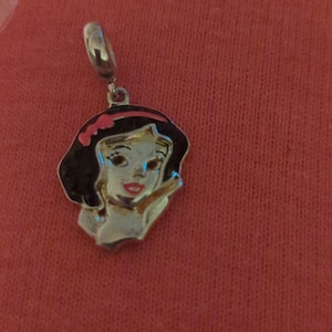 May include: A silver charm featuring Snow White from Disney's Snow White and the Seven Dwarfs. The charm has a red bow and a white background with a detailed image of Snow White's face.