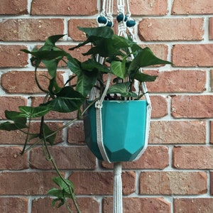May include: A teal ceramic planter with a leafy green plant hanging from a macrame plant hanger with white cord and teal beads. The hanger is attached to a brick wall.