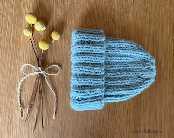 May include: A pale blue knitted beanie hat with a folded-up brim. The hat is made of a soft, ribbed knit material. Beside the hat is a small bouquet of yellow flowers tied with twine, all on a wooden surface.