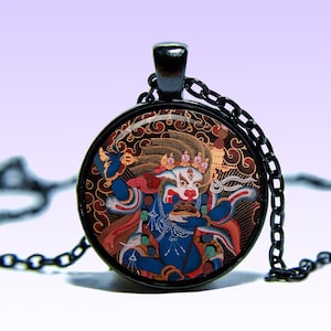 May include: A black chain necklace with a round glass pendant featuring a colorful illustration of a deity with a fierce expression. The deity is depicted with multiple arms and hands, holding various objects. The pendant is surrounded by a black metal frame.