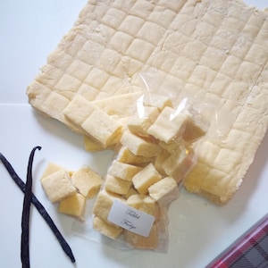 Könnte beinhalten: Draufsicht auf eine Charge Tablet Fudge, mit einer großen, geriffelten Platte und einem durchsichtigen Beutel mit geschnittenen Stücken. Zwei dunkle Vanilleschoten und Geschenkpapier mit Karomuster sind ebenfalls vorhanden. Der Beutel trägt die Aufschrift "Tablet Fudge".