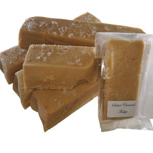 May include: A close-up of a stack of salted caramel fudge bars. The fudge is a light brown colour and has a grainy texture. The bars are wrapped in clear plastic and labelled "Salted Caramel Fudge".