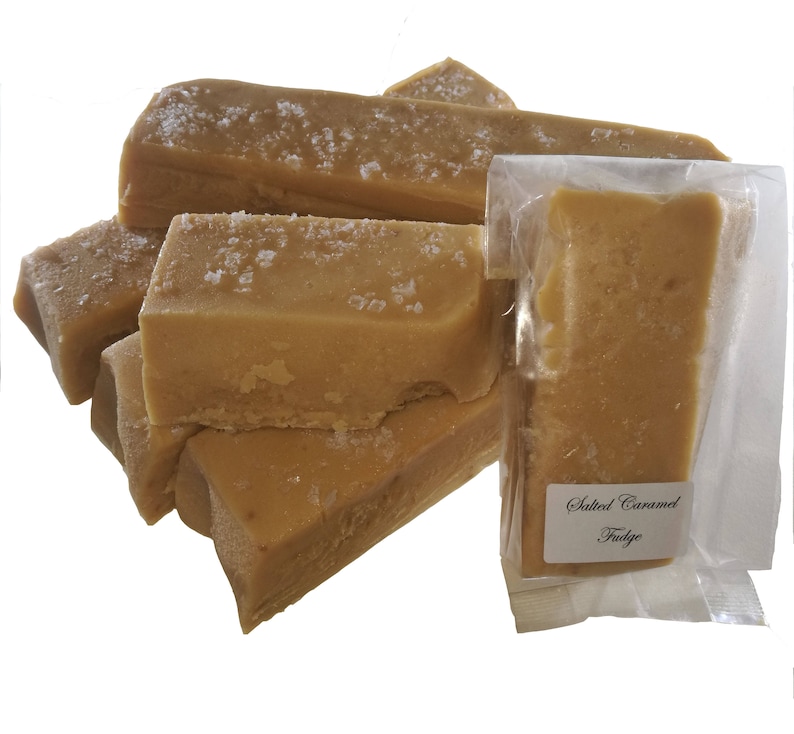 May include: A close-up of a stack of salted caramel fudge bars. The fudge is a light brown colour and has a grainy texture. The bars are wrapped in clear plastic and labelled "Salted Caramel Fudge".
