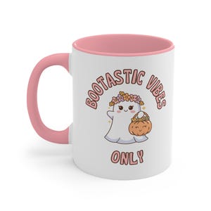 May include: A white ceramic mug with a pink handle and rim, featuring a cartoon ghost wearing a flower crown and holding a pumpkin basket. The text BOOTASTIC VIBES ONLY is printed in pink letters around the ghost, which has a smiling face.