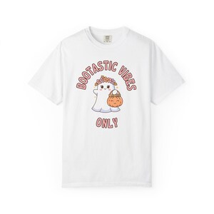May include: A white short-sleeve t-shirt featuring a graphic of a white ghost wearing a colorful flower crown and holding an orange jack-o'-lantern bucket. The text BOOTASTIC VIBES ONLY is printed in pink letters above and below the ghost illustration.