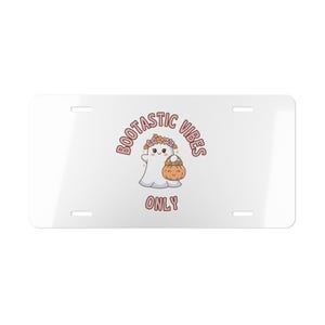 May include: A white rectangular license plate featuring a central graphic of a white ghost wearing a colorful flower crown and holding an orange pumpkin basket. The text BOOTASTIC VIBES ONLY is printed in pink letters above and below the ghost.
