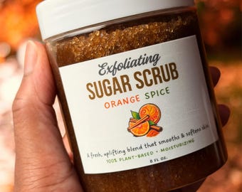 May include: A clear plastic jar filled with brown exfoliating sugar scrub, topped with a white lid. The label reads Exfoliating SUGAR SCRUB ORANGE SPICE, features an orange and cinnamon graphic, and states 8 FL OZ, 100% PLANT-BASED, MOISTURIZING.