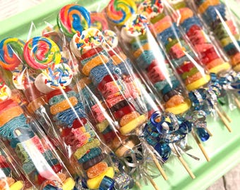 May include: A collection of candy skewers individually wrapped in clear plastic. Each skewer features a colorful lollipop, a marshmallow topped with rainbow sprinkles, and various gummy candies in red, orange, yellow, green, and blue.