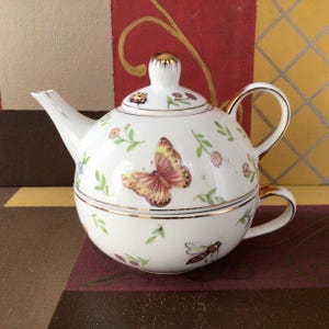 May include: A white porcelain tea-for-one set featuring a teapot stacked on a teacup. The set is decorated with colourful butterflies, a ladybug, a bee, and small floral sprigs. The teapot lid, spout, and handles are accented with gold trim.