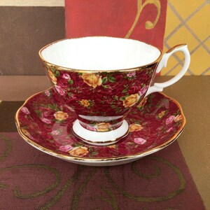 Può includere: Un set vintage di tazza e piattino da tè. La tazza e il piattino sono di un rosso intenso con un motivo floreale di rose gialle e rosa. I bordi sono rifiniti in oro. La tazza ha un interno bianco e un manico bianco.