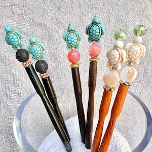 May include: Five wooden hair sticks with decorative tops. The tops feature turquoise and white sea turtles, pink and orange beads, and green and white beads. The sticks are displayed in a clear glass jar filled with white sand.
