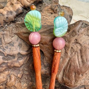 May include: Two wooden hair sticks with decorative tops. Each stick features a light green and blue seashell-shaped bead, a pink sphere, and a small gold bead accent. The sticks have a warm, reddish-brown hue.