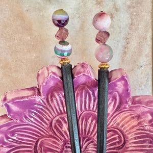 May include: Two dark wooden hair sticks featuring decorative tops with stacked beads. Each stick has a gold-toned metal spacer, a faceted translucent purple bead, and two round patterned stone beads in shades of pink, green, and white.