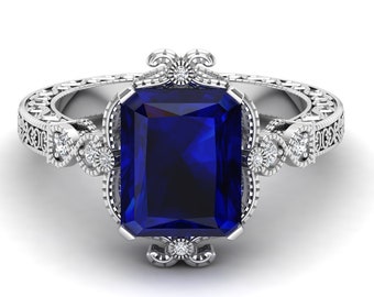 May include: A silver ring with a large, deep blue, emerald-cut sapphire gemstone surrounded by smaller diamonds. The ring has a delicate, ornate design.