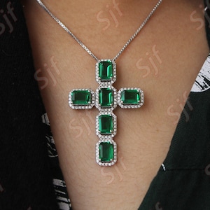 May include: A silver necklace with a cross pendant. The cross features emerald-cut green gemstones surrounded by small, clear stones. The necklace is worn against a dark background.