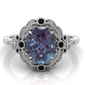 Puede incluir: Anillo de plata con una piedra preciosa de alejandrita de talla ovalada que cambia de color. El anillo presenta un engaste decorativo con detalles en negro y un dise&ntilde;o de filigrana detallado.