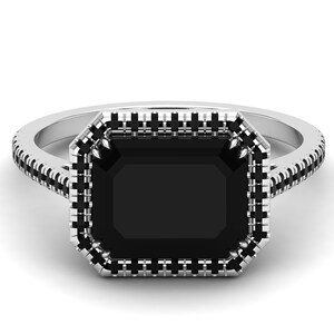 Op de afbeelding: Een zilveren ring met een grote, rechthoekige zwarte edelsteen. De edelsteen is omgeven door een halo van kleinere zwarte stenen, en de band is ook gedetailleerd met zwarte stenen. De ring staat tegen een witte achtergrond.