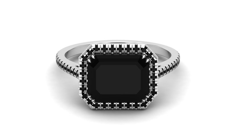 Op de afbeelding: Een zilveren ring met een grote, rechthoekige zwarte edelsteen. De edelsteen is omgeven door een halo van kleinere zwarte stenen, en de band is ook gedetailleerd met zwarte stenen. De ring staat tegen een witte achtergrond.