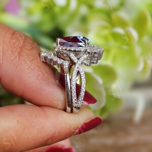 May include: A silver ring set with a large, oval-shaped, red gemstone surrounded by smaller, clear gemstones. The ring is set on a band with a twisted design and a smaller band with a row of clear gemstones.