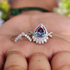 May include: A pear-shaped alexandrite engagement ring set with a halo of smaller diamonds. The ring is set in white gold and features a delicate, curved wedding band with small diamonds.
