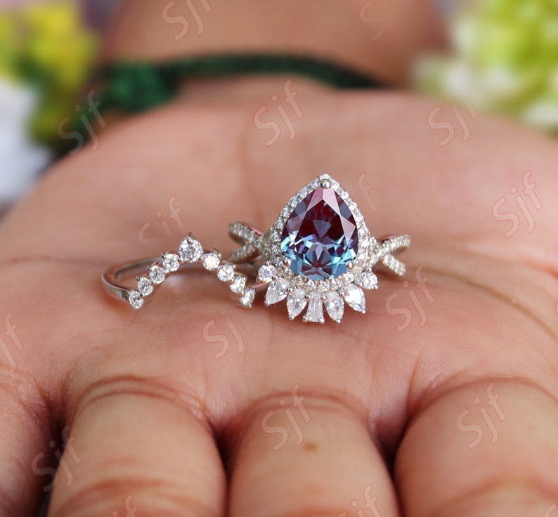 May include: A pear-shaped alexandrite engagement ring set with a halo of smaller diamonds. The ring is set in white gold and features a delicate, curved wedding band with small diamonds.