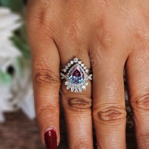 May include: A pear-shaped, blue and red gemstone engagement ring with a halo of smaller diamonds. The ring is set in silver and has a matching band with a delicate design.