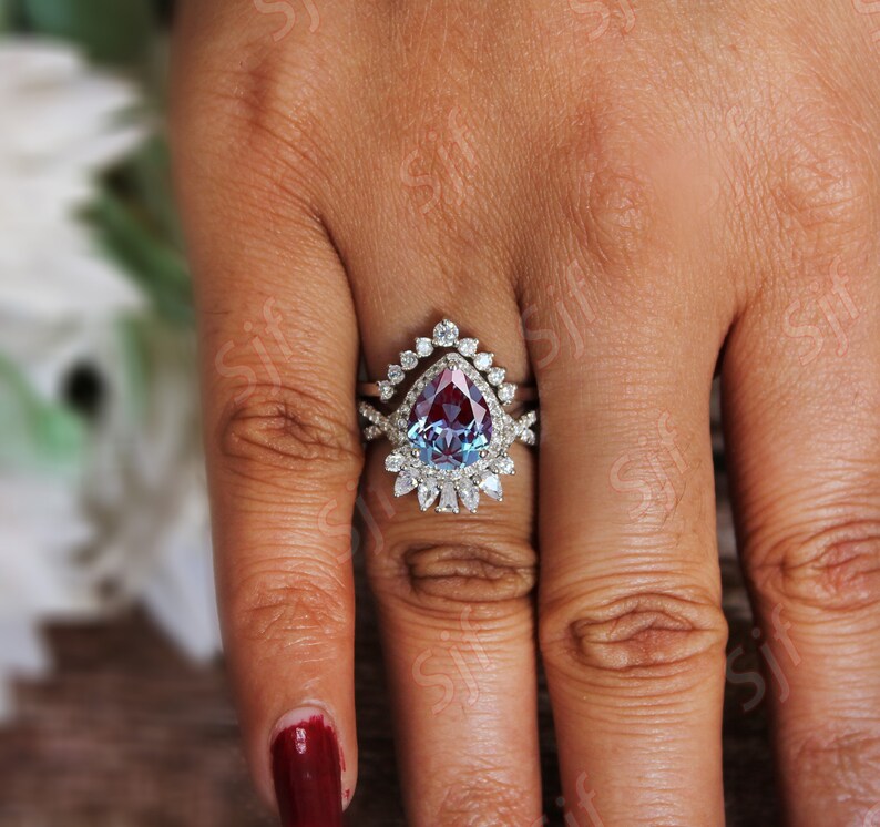 May include: A pear-shaped, blue and red gemstone engagement ring with a halo of smaller diamonds. The ring is set in silver and has a matching band with a delicate design.