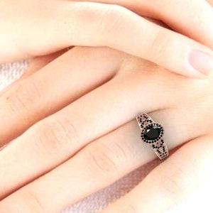 May include: A rose gold ring featuring a black oval gemstone surrounded by smaller black gemstones. The ring is worn on a finger.