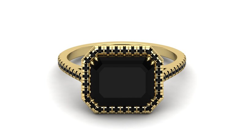 Op de afbeelding: Een gouden ring met een grote, rechthoekige zwarte edelsteen. De ring heeft een halo van kleinere zwarte stenen rond de centrale steen en langs de band.