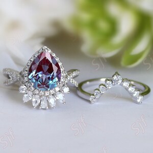 May include: A pear-shaped, iridescent gemstone ring with a halo of smaller diamonds. The ring is set in silver and has a delicate, twisted band. A matching, curved band with smaller diamonds is also shown.