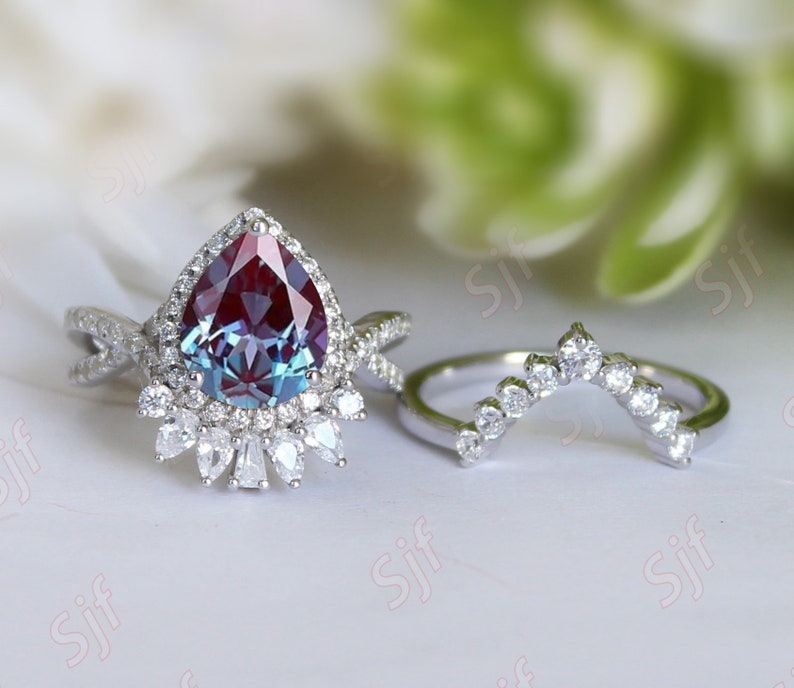 May include: A pear-shaped, iridescent gemstone ring with a halo of smaller diamonds. The ring is set in silver and has a delicate, twisted band. A matching, curved band with smaller diamonds is also shown.