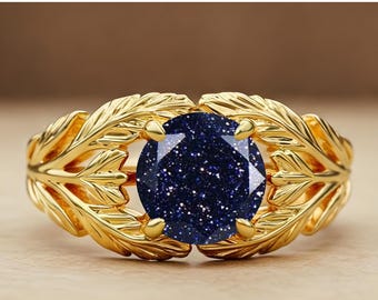 Könnte beinhalten: Ein goldfarbener Ring mit einem großen, runden, dunkelblauen Edelstein mit funkelnden Einschlüssen. Der Ring hat ein Band mit aufwendigen blattartigen Designs, die eine elegante, von der Natur inspirierte Ästhetik schaffen. Der Edelstein wird von vier Krappen gehalten.