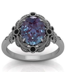 Puede incluir: Un anillo plateado con una piedra preciosa ovalada que cambia de color. El anillo presenta un dise&ntilde;o floral ornamentado alrededor de la piedra central, con peque&ntilde;os detalles negros. La banda tiene un patr&oacute;n texturizado detallado.