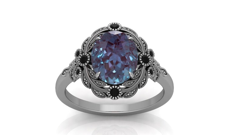 Puede incluir: Un anillo plateado con una piedra preciosa ovalada que cambia de color. El anillo presenta un dise&ntilde;o floral ornamentado alrededor de la piedra central, con peque&ntilde;os detalles negros. La banda tiene un patr&oacute;n texturizado detallado.