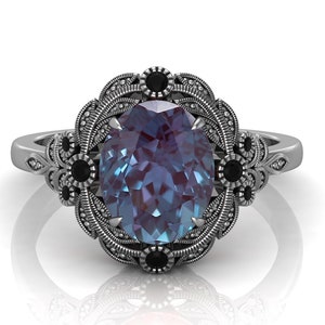 Puede incluir: Un anillo plateado con una piedra preciosa de alejandrita ovalada que cambia de color. El anillo tiene un engaste ornamentado, de inspiraci&oacute;n floral, con detalles en negro. La banda est&aacute; detallada con patrones delicados, creando una est&eacute;tica vintage.