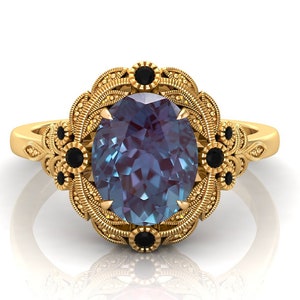 Puede incluir: Anillo dorado con una piedra preciosa ovalada de color azul-p&uacute;rpura. El anillo presenta un dise&ntilde;o floral ornamentado con detalles negros. La banda y el engaste est&aacute;n decorados con intrincados patrones.