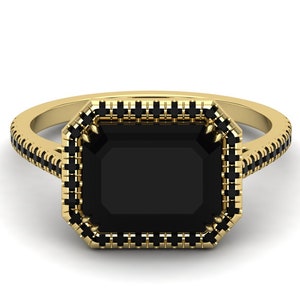 Op de afbeelding: Een gouden ring met een grote, rechthoekige zwarte edelsteen. De ring heeft een halo van kleinere zwarte stenen rond de centrale steen en langs de band, wat een opvallend contrast cre&euml;ert.