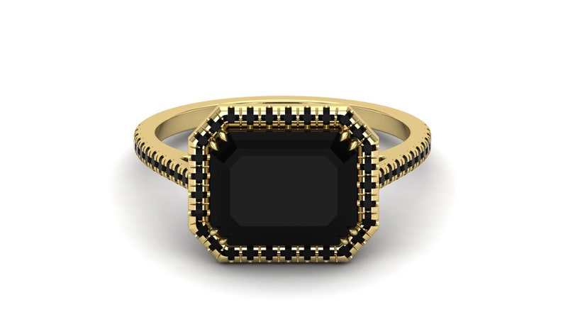 Op de afbeelding: Een gouden ring met een grote, rechthoekige zwarte edelsteen. De ring heeft een halo van kleinere zwarte stenen rond de centrale steen en langs de band, wat een opvallend contrast cre&euml;ert.