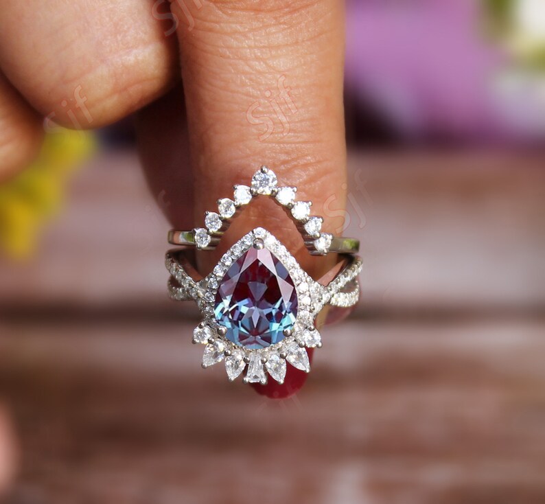 May include: A pear-shaped alexandrite engagement ring set with a halo of smaller diamonds. The ring is set in silver and has a delicate, intricate design.