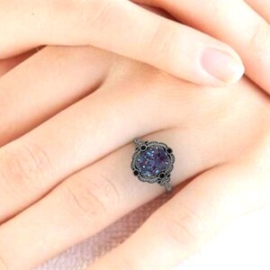 Puede incluir: Un anillo de metal oscuro con una gran piedra preciosa redonda de color p&uacute;rpura-azul en el centro. El anillo tiene un dise&ntilde;o calado decorativo alrededor de la piedra central y piedras negras m&aacute;s peque&ntilde;as. El anillo est&aacute; en el dedo de una persona.