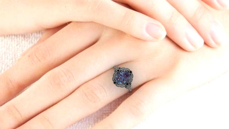Puede incluir: Un anillo de metal oscuro con una gran piedra preciosa redonda de color p&uacute;rpura-azul en el centro. El anillo tiene un dise&ntilde;o calado decorativo alrededor de la piedra central y piedras negras m&aacute;s peque&ntilde;as. El anillo est&aacute; en el dedo de una persona.