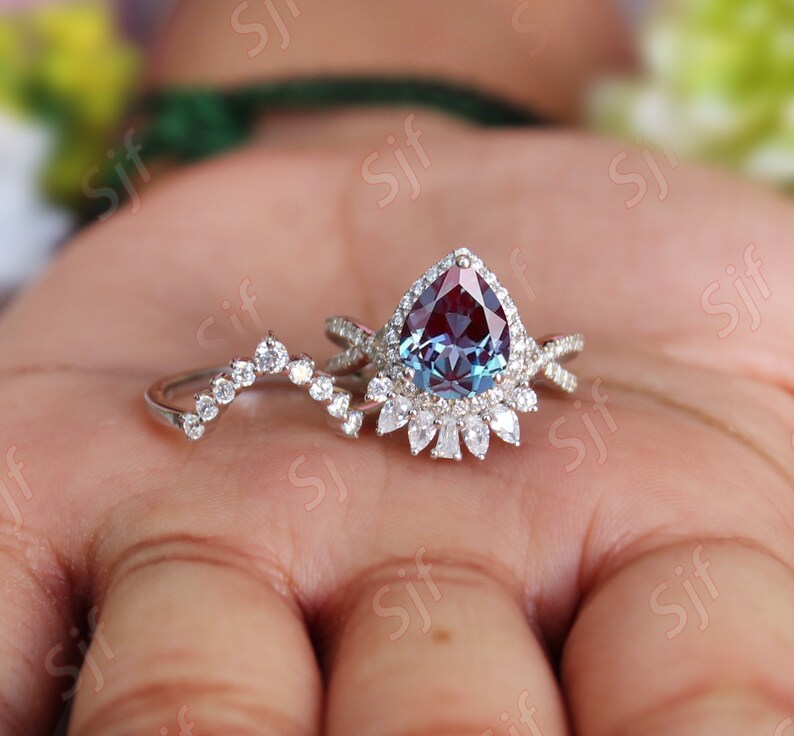 May include: A pear-shaped alexandrite engagement ring set with a halo of smaller diamonds. The ring is set in white gold and features a delicate, curved wedding band with small diamonds.