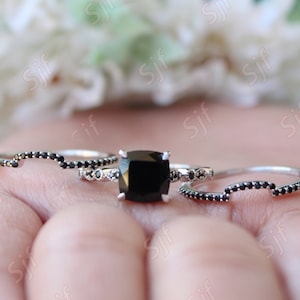 May include: A set of three black diamond engagement rings. The center ring features a large square-cut black diamond surrounded by smaller diamonds. The two side rings are thin bands with black diamonds set in a pave setting.