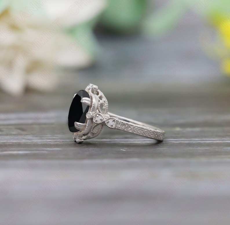 May include: A silver engagement ring with a black oval gemstone and intricate filigree details. The ring is set on a wooden surface.