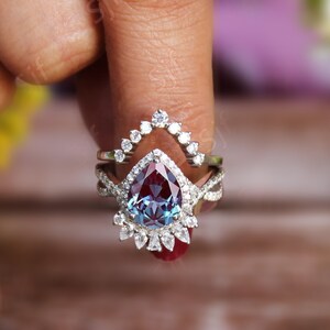May include: A pear-shaped alexandrite engagement ring set with a halo of smaller diamonds. The ring is set in silver and has a delicate, intricate design.