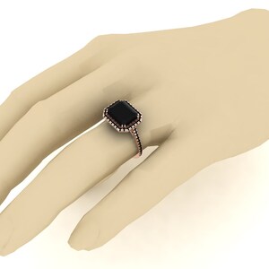 Op de afbeelding: Een ros&eacute;gouden ring met een grote, rechthoekige zwarte edelsteen. De edelsteen is omgeven door een halo van kleinere stenen. De ring wordt op een hand getoond.