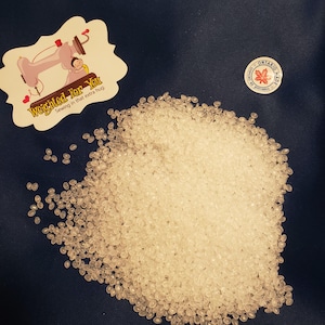 May include: A close-up of a pile of small, white, translucent plastic pellets. The pellets are oval-shaped and have a smooth, glossy finish.