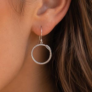 May include: A silver-toned earring featuring a circular snake design, known as an Ouroboros, suspended from a simple hook. The snake is depicted biting its own tail, forming a continuous ring shape with a textured, scaled surface finish.
